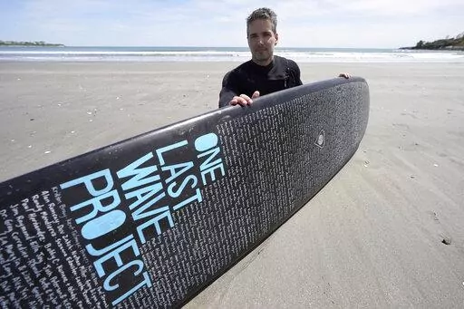 Dan Fischer, of Newport, R.I., sits for a photograph with his surfboard on Easton's Beach, in Newport, Wednesday, May 18, 2022. Fischer, 42, created the One Last Wave Project in January 2022 to use the healing power of the ocean to help families coping with a loss, as it helped him following the death of his father. Fischer places names onto his surfboards, then takes the surfboards out into the ocean as a way to memorialize the lost loved ones in a place that was meaningful to them. (AP Photo
