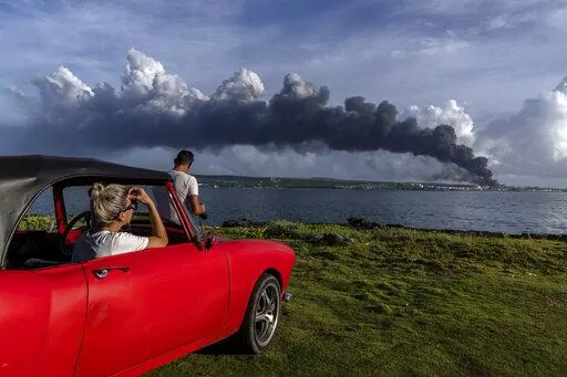 People watch a huge plume of smoke rise from the Matanzas supertanker base, as firefighters work to douse a fire that started during a thunderstorm the night before, in Matanzas, Cuba, Sunday, Aug. 7, 2022. Cuban authorities say lightning struck a crude oil storage tank at the base, sparking a fire that sparked four explosions that injured more than 121 people, one person dead and 17 missing. (AP Photo/Ramon Espinosa)