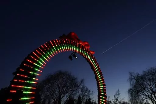 A commercial air plane streaks across the the post-sunset sky above the "Welcome Arch" at the beginning of a 1.3-mile path through the Chicago Botanic Garden's fifth annual Lightscape holiday experience of light and music in Glencoe, Ill., on Thursday, Dec. 14, 2023. (AP Photo/Charles Rex Arbogast)