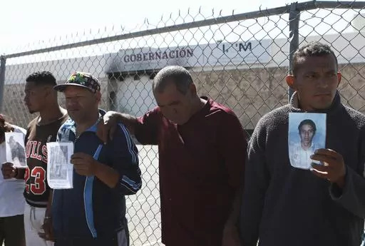 Migrants, mostly from Venezuela, hold photos of those who died in a fire at a Mexican immigration detention center, behind, during a prayer vigil outside the center in Ciudad Juarez, Mexico, April, 27, 2023. Four months after a fire in a Mexican immigration detention center at the border killed 40 migrants, some survivors are living in limbo at a Mexico City hotel, recovering from their injuries and awaiting the prosecution of their captors. (AP Photo/Christian Chavez, File)