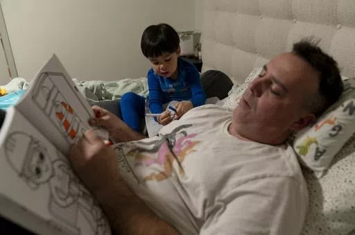 Scott Quinn and his 4-year-old son, Ethan, work on coloring books in a bed in Concord, Calif., Wednesday, Nov. 1, 2023. Ethan's parents are among those who have opted for a private daycare instead of a free “transitional kindergarten.” Quinn said he has been discouraged to see Ethan — one of the oldest kids in his daycare class — pick up the behavior of kids who are several years younger than him. “In retrospect, it would have been better to send him to school to be around kids his age