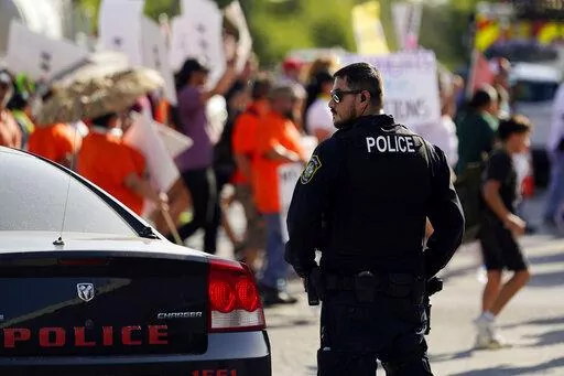 A Uvalde police officer watches as family and friends of those killed and injured in the school shootings at Robb Elementary take part in a protest march and rally, Sunday, July 10, 2022, in Uvalde, Texas.   Four months after the Robb Elementary School shooting, the Uvalde school district on Friday, Oct. 7 pulled its entire embattled campus police force off the job following a wave of new outrage over the hiring of a former Texas state trooper who was part of the hesitant law enforcement respons