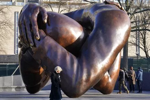 Passers-by walk near the 20-foot-high bronze sculpture "The Embrace," a memorial to Dr. Martin Luther King Jr. and Coretta Scott King, in the Boston Common, Tuesday, Jan. 10, 2023, in Boston. The sculpture, consisting of four intertwined arms, was inspired by a photo of the Kings embracing when MLK learned he had won the Nobel Peace Prize in 1964. The statue is to be unveiled during ceremonies Friday, Jan. 13, 2023. (AP Photo/Steven Senne)