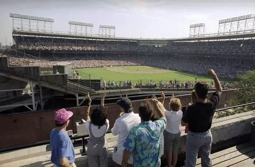 In this Monday, July 9, 1990, file photo, spectators watch an All-Star Game practice session from the roof of a building just outside Chicago's Wrigley Field. Booking hotels for baseball games during shoulder seasons like May, June or September can often be cheaper than the summer months. Additionally, save money by planning to attend weekday versus weekend games. (AP Photo/Seth Perlman, File)